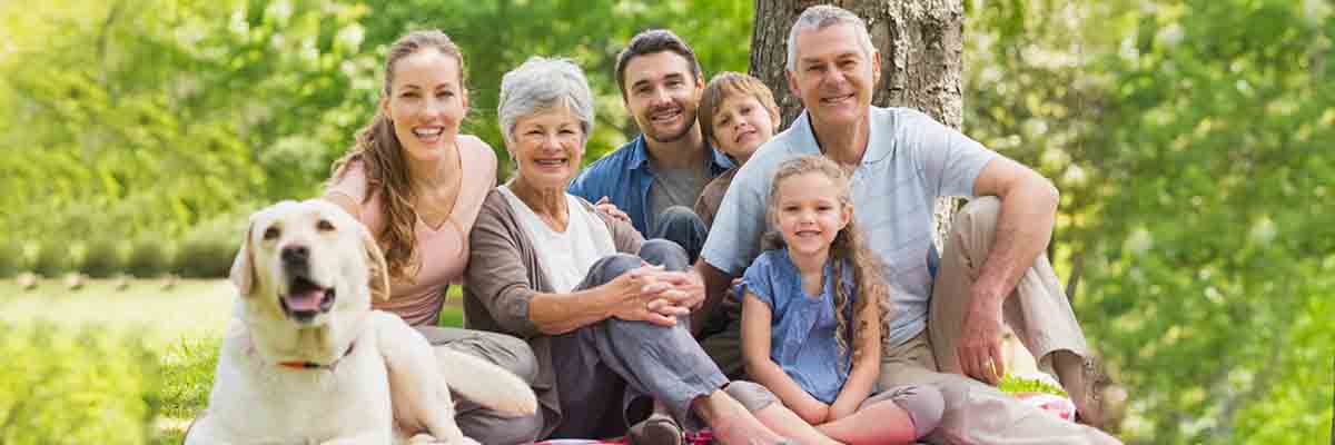 A family of four sits on a blanket in a park, smiling, with a dog lying beside them
